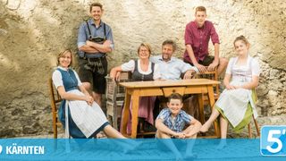 Eine Familie von sieben Personen, die lächelnd und in traditioneller Kleidung für ein Foto posiert, wobei zwei am Tisch sitzen und fünf stehend dahinter.