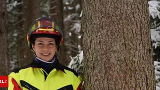 Eine Frau in einer reflektierenden Sicherheitsjacke mit Helm steht neben einem Baum in einem verschneiten Wald.