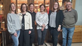 Fünf Frauen stehen zusammen und lächeln für ein Foto. Sie tragen Freizeitkleidung und Brillen. Die Frau in der Mitte hat eine Halskette. Hinter ihnen ist ein hölzerner Schrank mit Glastüren und Regalen zu sehen.