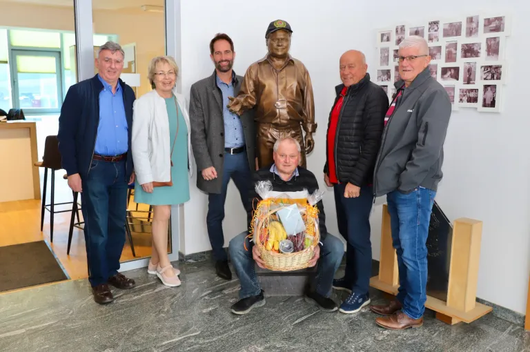Sieben Personen stehen vor einer Statue und posieren für ein Foto. Ein Mann kniet mit einem Korb voller Früchte. Die Statue trägt Hut und Uniform. Hinter ihnen befindet sich eine Fotocollage an der Wand.