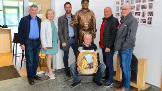 Sieben Personen stehen vor einer Statue und posieren für ein Foto. Ein Mann kniet mit einem Korb voller Früchte. Die Statue trägt Hut und Uniform. Hinter ihnen befindet sich eine Fotocollage an der Wand.
