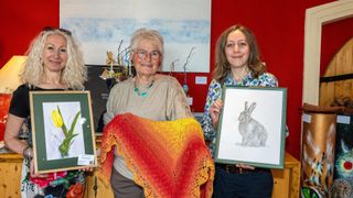 Drei Frauen präsentieren Kunstwerke; eine hält ein gerahmtes Kaninchen-Zeichnung, eine andere ein gestricktes Tuch, und die dritte ein gerahmtes Bild. Sie stehen vor einer roten Wand mit Kunst und Grünpflanzen.