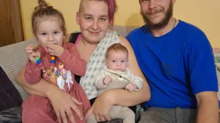 Eine lächelnde Familie sitzt auf einem Sofa. Eine Frau mit rosa Haaren hält ein Baby und ein Mann mit Bart hält ein Kleinkind. Das Kleinkind hält ein Spielzeug.
