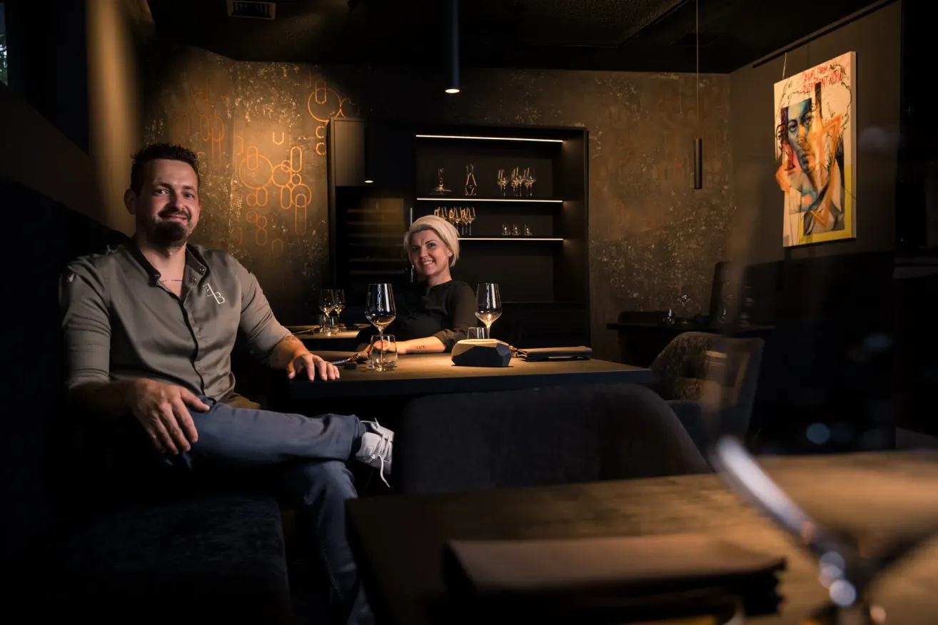 Ein Mann und eine Frau sitzen an einem Tisch in einem Restaurant, lächeln und posieren für ein Foto. Hinter ihnen sind Regale mit Weingläsern und Flaschen.