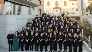 Ein Gruppenfoto eines Musikensembles in schwarzer Kleidung, das verschiedene Instrumente wie Trompeten und Flöten hält, steht auf einer Treppe vor einem Gebäude.