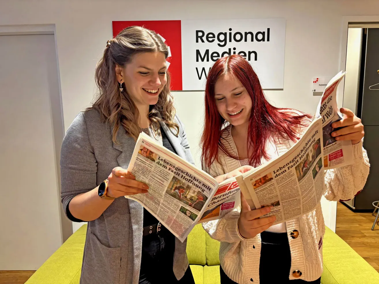 Zwei junge Frauen mit langen Haaren lächeln, während sie Zeitungen lesen. Die Frau auf der linken Seite trägt eine graue Strickjacke, und die Frau auf der rechten Seite trägt eine weiße Strickjacke. Sie stehen in der Nähe einer Wand mit dem Text 'Regional Medien W'.