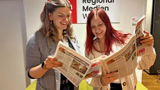 Zwei junge Frauen mit langen Haaren lächeln, während sie Zeitungen lesen. Die Frau auf der linken Seite trägt eine graue Strickjacke, und die Frau auf der rechten Seite trägt eine weiße Strickjacke. Sie stehen in der Nähe einer Wand mit dem Text 'Regional Medien W'.