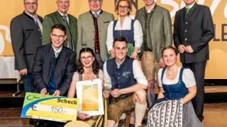 Eine Gruppe von Menschen in traditioneller bayerischer Tracht posiert für ein Foto, einige halten ein Zertifikat und einen Scheck. Hinter ihnen ist ein Banner mit der Aufschrift 'FIN B' zu sehen.