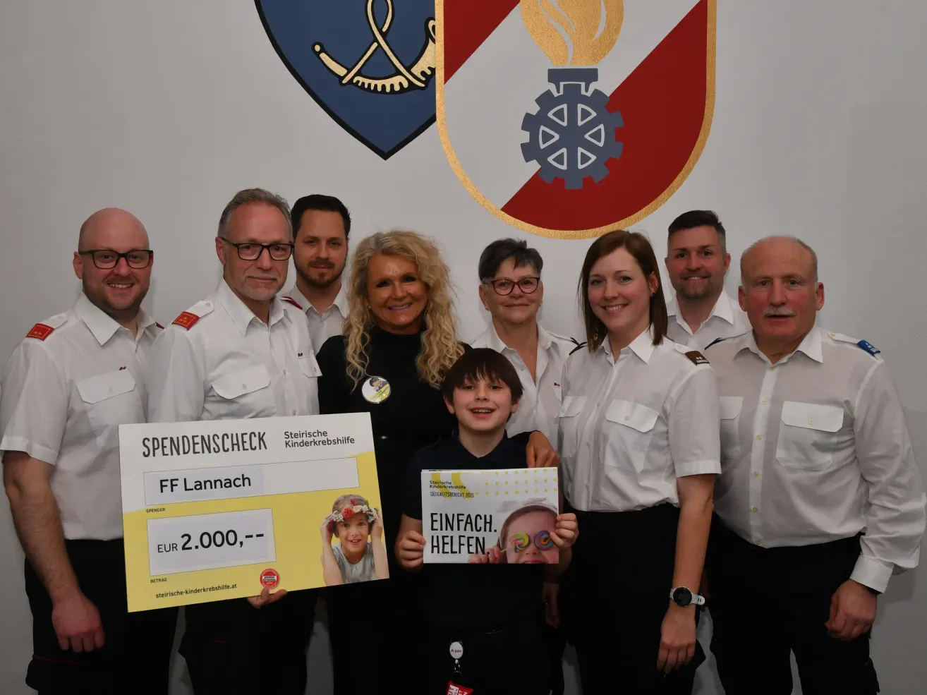 Eine Gruppe von Erwachsenen und Kindern posiert für ein Foto mit einem Spendenscheck. Ein Junge hält einen Scheck mit einem bemalten Gesicht. Dahinter befindet sich ein Schild mit einem Logo.
