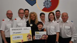 Eine Gruppe von Erwachsenen und Kindern posiert für ein Foto mit einem Spendenscheck. Ein Junge hält einen Scheck mit einem bemalten Gesicht. Dahinter befindet sich ein Schild mit einem Logo.