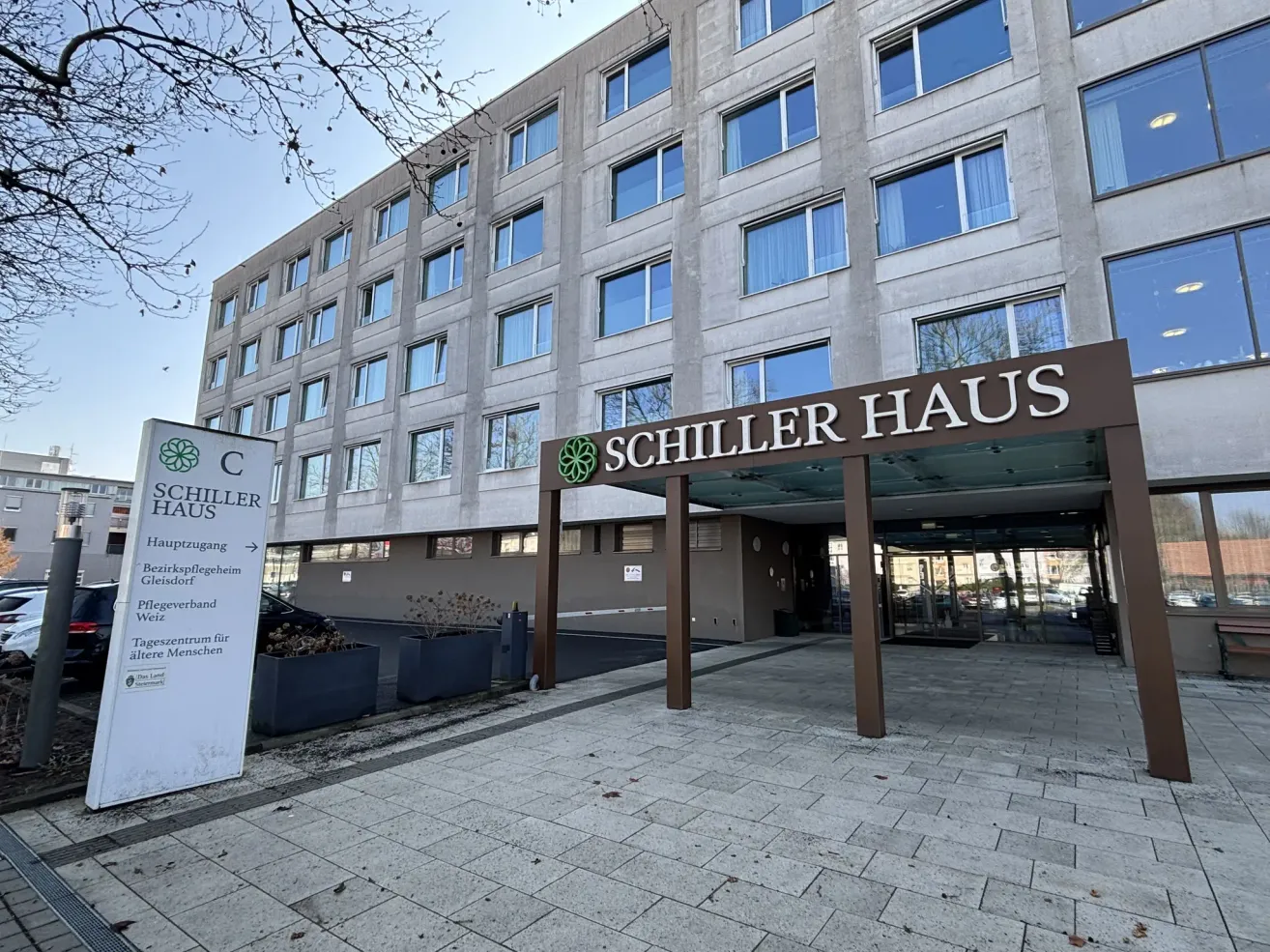 Eingang des Schiller Haus mit großen Fenstern, einem Schild mit einem grünen Logo und einem gepflasterten Weg davor.