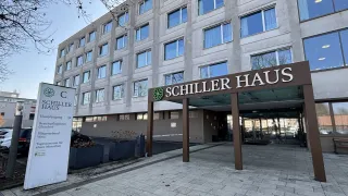 Eingang des Schiller Haus mit großen Fenstern, einem Schild mit einem grünen Logo und einem gepflasterten Weg davor.