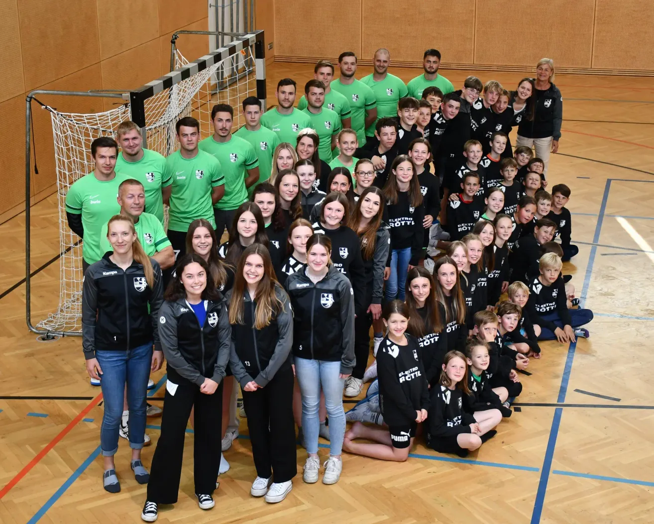 Eine große Gruppe von Menschen in grünen und schwarzen Trikots steht in einer Sporthalle und posiert für ein Gruppenfoto mit einem Netz und einem weißen Boden.