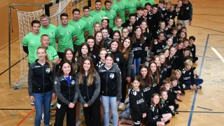 Eine große Gruppe von Menschen in grünen und schwarzen Trikots steht in einer Sporthalle und posiert für ein Gruppenfoto mit einem Netz und einem weißen Boden.
