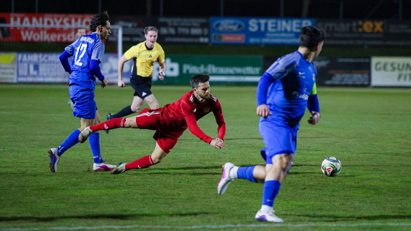 Fußballspieler in Aktion auf einem Feld bei Nacht. Ein Spieler in Rot taucht, während ein anderer in Blau auf ihn zuläuft. Ein Offizieller beobachtet von hinten.