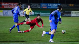 Fußballspieler in Aktion auf einem Feld bei Nacht. Ein Spieler in Rot taucht, während ein anderer in Blau auf ihn zuläuft. Ein Offizieller beobachtet von hinten.