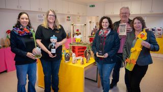 Vier Erwachsene stehen um einen Tisch mit Osterdekoration. Die Frau auf der linken Seite hält eine Flasche. Die Frau auf der rechten Seite hält eine Box. Die beiden Männer stehen an der Seite.