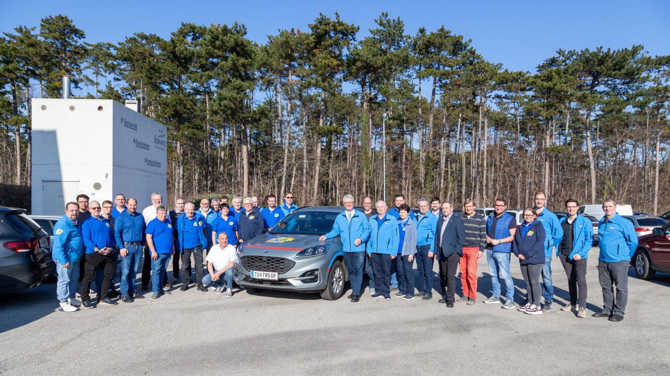 Eine Gruppe von Menschen in blauen Jacken posiert mit einem grauen Auto in einem Waldgebiet. Bäume stehen hinter ihnen.