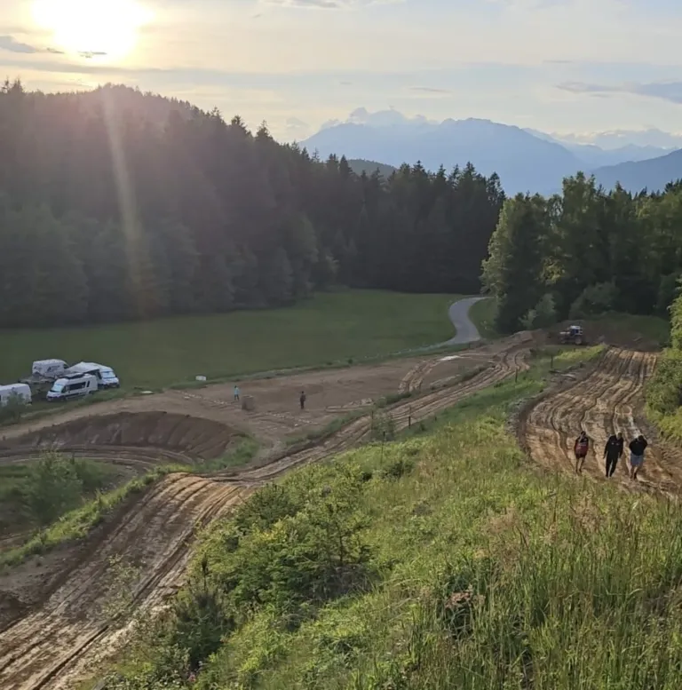 Eine Gruppe von Menschen geht auf einem unbefestigten Pfad in einem ländlichen Gebiet mit Bergen und einem Feld im Hintergrund. Fahrzeuge sind an der Seite geparkt.