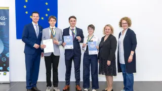 Sechs Personen stehen vor einer Europaflagge. Zwei Männer und eine Frau halten Urkunden und Medaillen. Die Frau trägt eine Brille, und die Männer tragen Anzüge und Krawatten. Die Frau auf der rechten Seite trägt eine Armbanduhr.