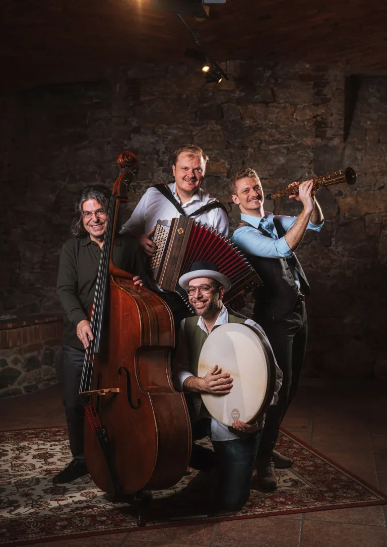 Vier Männer mit Instrumenten posieren für ein Foto. Der Mann in der Mitte spielt eine Trommel. Der Mann auf der rechten Seite spielt Klarinette. Der Mann auf der linken Seite spielt Cello. Der Mann in der Mitte spielt Akkordeon.