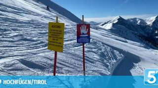 Ein gelbes Schild mit 'ACHTUNG!' und weiterem Text steht neben einem roten Schild mit Handsymbol, beide auf Stangen auf einem verschneiten Berg.