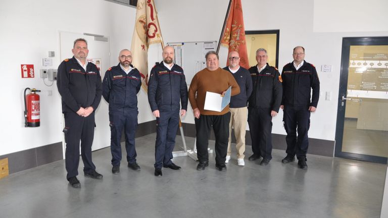 Sechs Männer stehen in einem Raum mit weißen Wänden, tragen Uniformen, halten Papiere und stehen vor Flaggen.