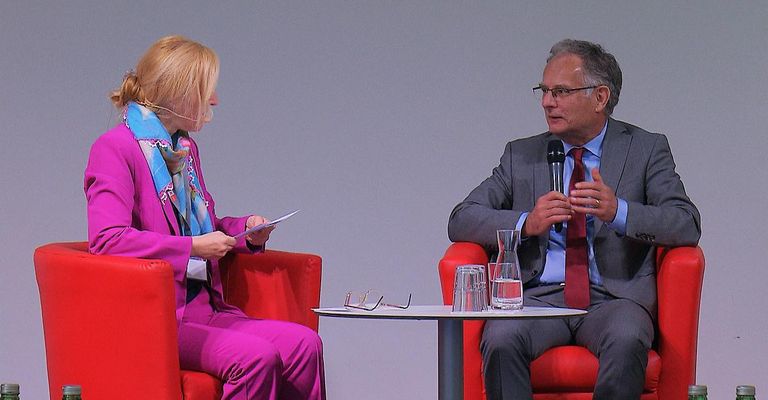 Eine Frau und ein Mann sitzen in roten Stühlen und sehen sich gegenüber. Der Mann hält ein Mikrofon. Die Frau hält ein Papier und einen Stift. Ein Tisch zwischen ihnen hat zwei Gläser und eine Vase.