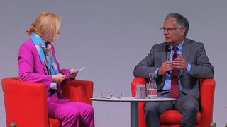 Eine Frau und ein Mann sitzen in roten Stühlen und sehen sich gegenüber. Der Mann hält ein Mikrofon. Die Frau hält ein Papier und einen Stift. Ein Tisch zwischen ihnen hat zwei Gläser und eine Vase.