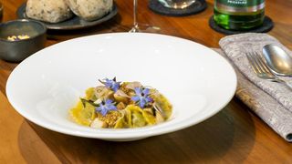 Eine weiße Schüssel mit Knödeln und blauen Blumen auf einem Holztisch, begleitet von einem Weinglas und einer Flasche Prickeln.