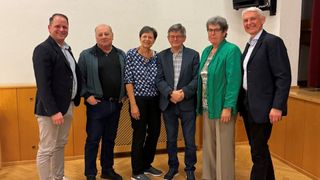 Fünf Menschen stehen zusammen in einem Raum mit Holzböden. Sie lächeln alle und scheinen für ein Foto zu posieren. Sie sind alle formell gekleidet.