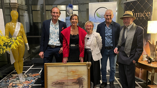 Fünf Personen posieren für ein Foto, stehend um ein gerahmtes Gemälde herum. Das Gemälde zeigt eine Landschaft. Hinter ihnen befindet sich eine Glaswand und eine Puppe. Im Hintergrund sind Banner sichtbar.