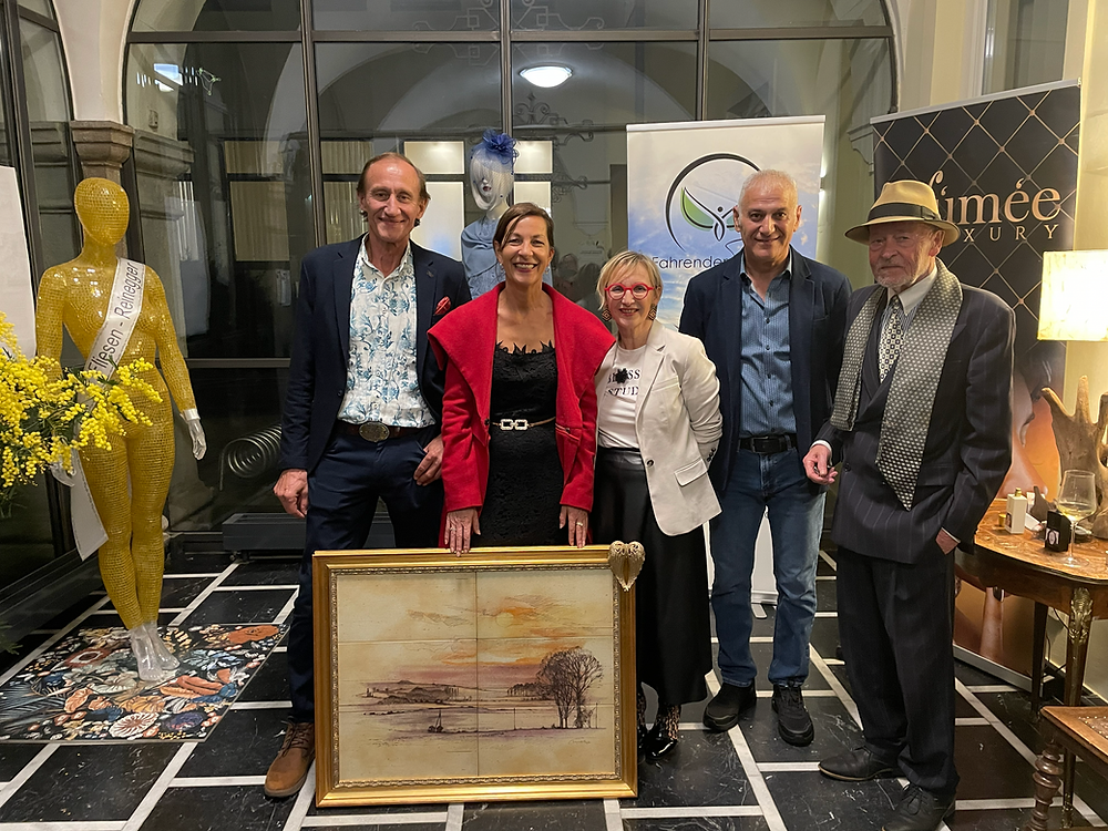 Fünf Personen posieren für ein Foto, stehend um ein gerahmtes Gemälde herum. Das Gemälde zeigt eine Landschaft. Hinter ihnen befindet sich eine Glaswand und eine Puppe. Im Hintergrund sind Banner sichtbar.