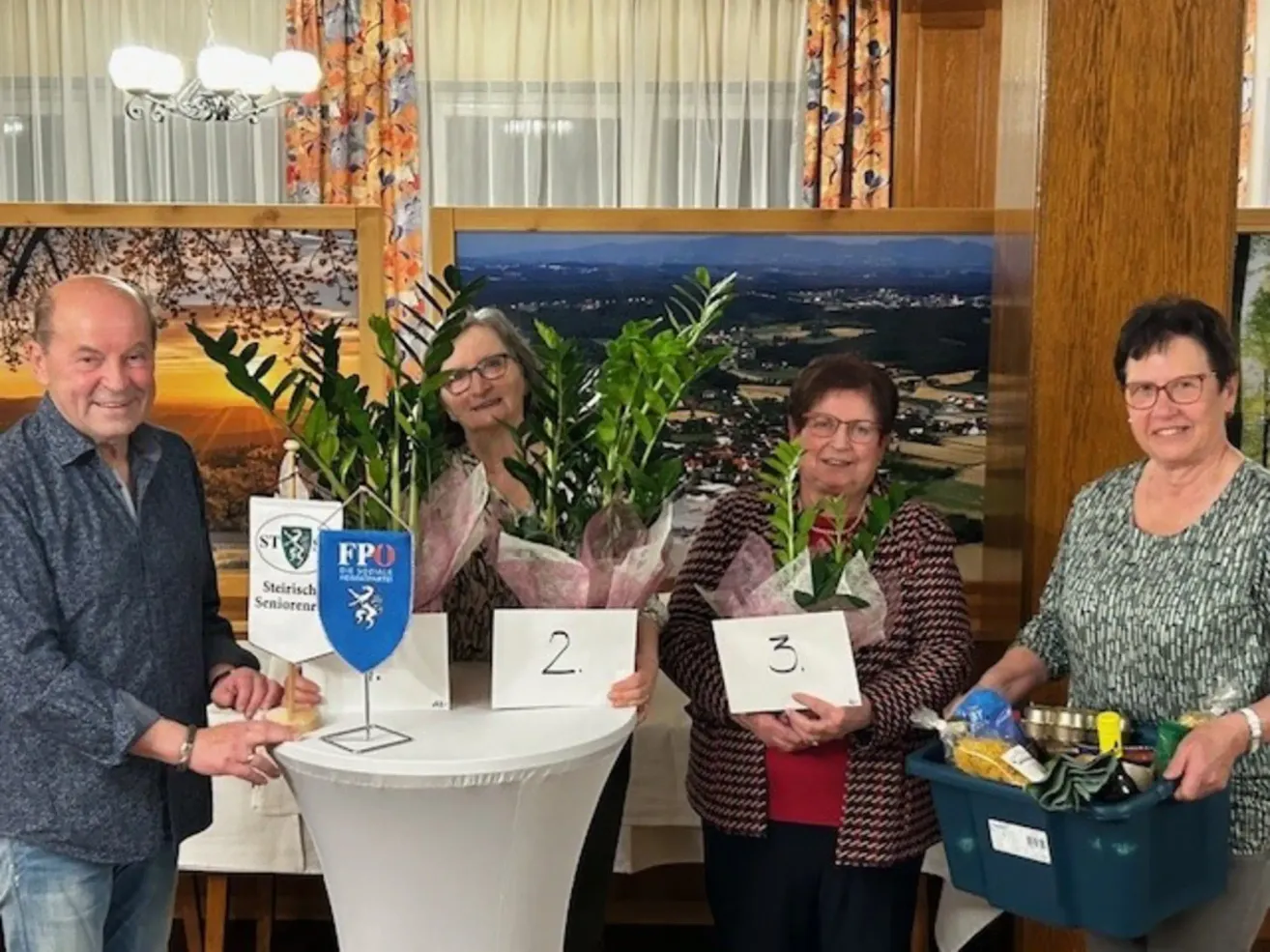 Vier Frauen stehen hinter einem Tisch und halten Blumen und Schilder. Sie lächeln und posieren für ein Foto. Ein blauer Kasten mit Gegenständen ist rechts.