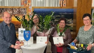 Vier Frauen stehen hinter einem Tisch und halten Blumen und Schilder. Sie lächeln und posieren für ein Foto. Ein blauer Kasten mit Gegenständen ist rechts.