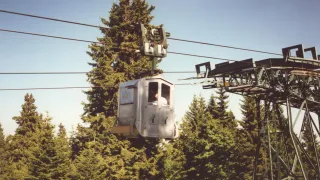 Eine Seilbahnkabine hängt von Seilen über einem Wald mit einer Person im Inneren, unter einem klaren Himmel.