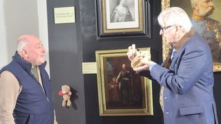 Ein Mann in einem blauen Anzug hält ein Sandwich vor ein Gemälde eines Mannes in Uniform. Ein Teddybär hängt an der Wand neben einem Informationsschild.