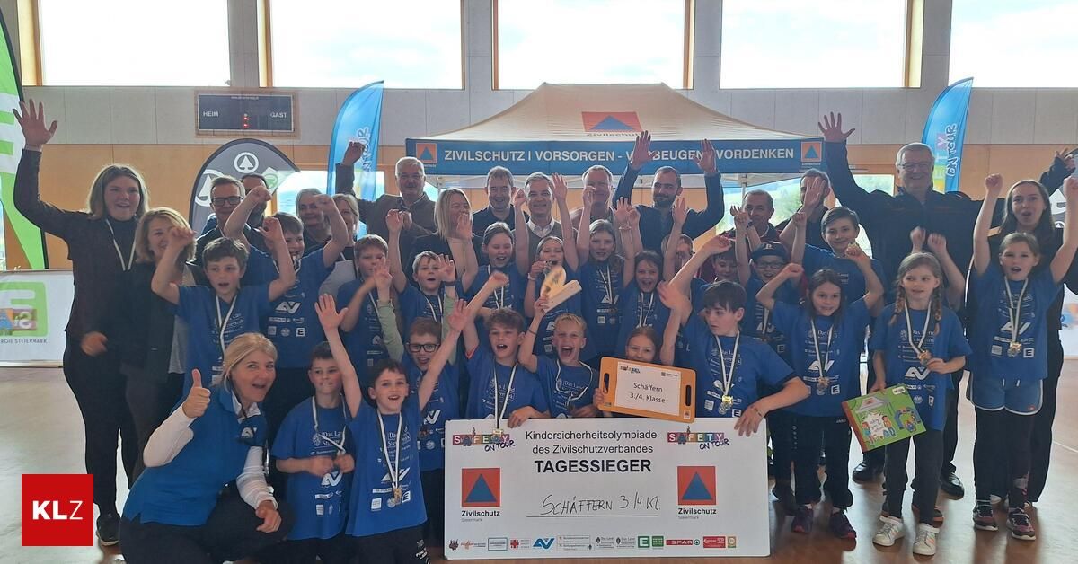 Eine Gruppe von Kindern und Erwachsenen in passenden blauen Hemden posiert für ein Foto und hält ein großes Schild hoch. Das Schild trägt die Aufschrift 'Kindersicherheitsolympiade des Zivilschutzverbandes'. Die Kinder haben Medaillen um den Hals und wirken aufgeregt. Sie befinden sich in einem Raum mit Holzboden und Fenstern.