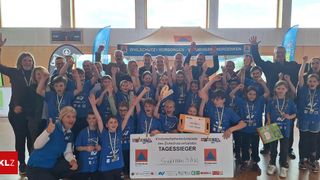 Eine Gruppe von Kindern und Erwachsenen in passenden blauen Hemden posiert für ein Foto und hält ein großes Schild hoch. Das Schild trägt die Aufschrift 'Kindersicherheitsolympiade des Zivilschutzverbandes'. Die Kinder haben Medaillen um den Hals und wirken aufgeregt. Sie befinden sich in einem Raum mit Holzboden und Fenstern.