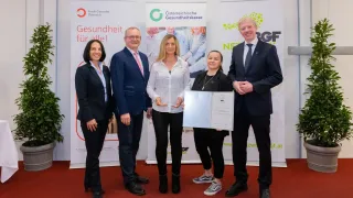 Fünf Personen stehen auf einem roten Teppich und lächeln. Eine Frau in der Mitte hält ein Zertifikat, mit zwei Männern auf beiden Seiten. Hinter ihnen sind Banner mit Logos und Text.