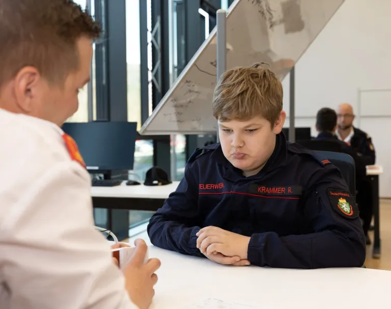 Zwei junge Jungen in Uniform sitzen an einem Schreibtisch in einem Büro. Ein Junge schaut den anderen an. Auf dem Schreibtisch steht eine Tasse.
