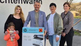 Eine Gruppe von Menschen posiert mit einer Box eines HP-Druckers. Der Mann hält die Box mit beiden Händen, und die Frau neben ihm lächelt. Dahinter ist eine Wand mit hebräischem Text.