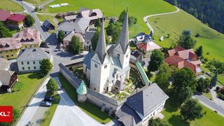 Ein Luftbild einer historischen Kirche mit zwei Türmen, umgeben von einem grünen Rasen und Häusern. Bäume und Autos sind im Hintergrund zu sehen.