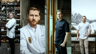 Ein Koch mit Bart und weißer Kochjacke und ein anderer Mann in einem schwarzen T-Shirt, der neben einem Fenster steht.