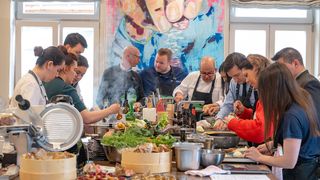Eine Gruppe von Menschen kocht gemeinsam in einer Küche, mit einem Koch vor ihnen. Es gibt mehrere Gerichte, Flaschen und Utensilien auf dem Tisch. Ein farbenfrohes Gemälde hängt an der Wand hinter ihnen.