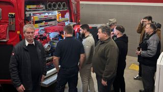 Eine Gruppe von Männern in unterschiedlicher Kleidung versammelt sich um einen roten Feuerwehrwagen. Einige inspizieren die Ausrüstung.