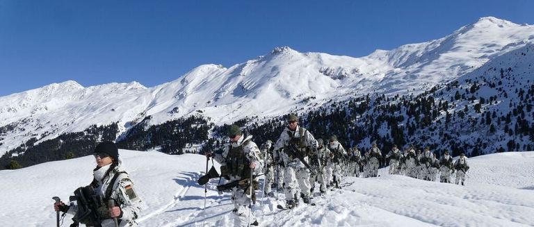 Soldaten in weißen Uniformen marschieren im Schnee, ausgestattet mit Skiern und Gewehren. Die Berge im Hintergrund sind schneebedeckt.