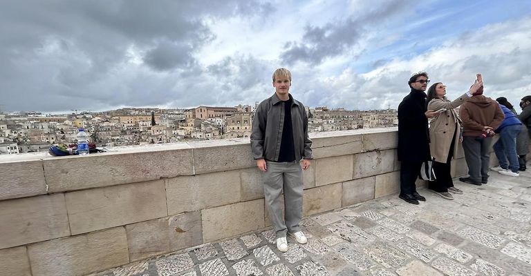 Ein Mann steht auf einer Steinmauer mit Blick auf eine Stadtlandschaft unter einem bewölkten Himmel. Er trägt eine graue Jacke und weiße Turnschuhe. Neben ihm steht eine weitere Person mit Sonnenbrille.