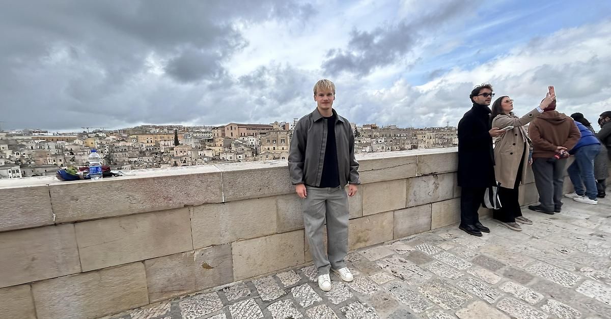 Ein Mann steht auf einer Steinmauer mit Blick auf eine Stadtlandschaft unter einem bewölkten Himmel. Er trägt eine graue Jacke und weiße Turnschuhe. Neben ihm steht eine weitere Person mit Sonnenbrille.