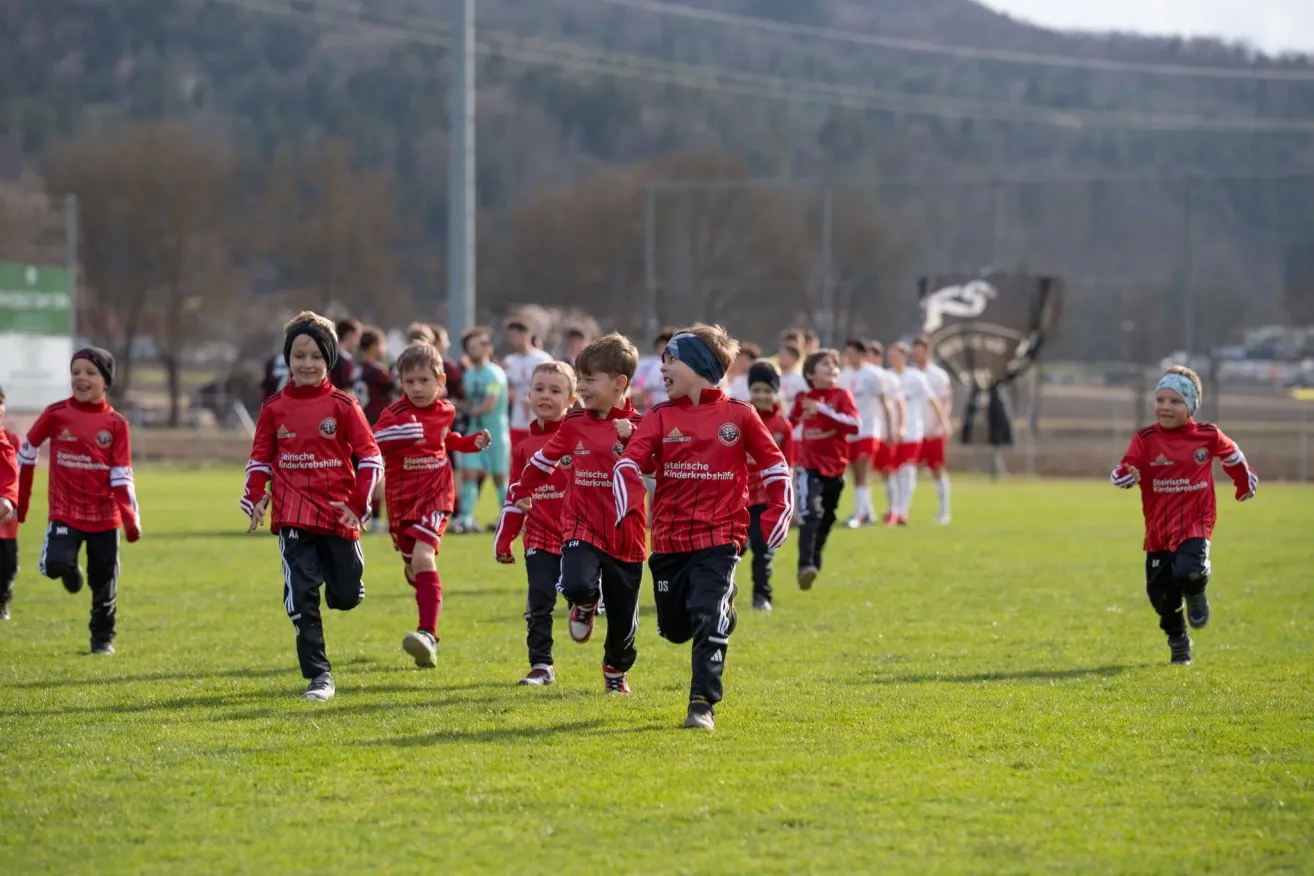 Eine Gruppe junger Jungen in roten Uniformen läuft auf einem grünen Feld, möglicherweise bei einem Fußballspiel. Sie werden von anderen Kindern und Erwachsenen verfolgt. In der Ferne befindet sich ein Pfahl mit Drähten und einem Berg.